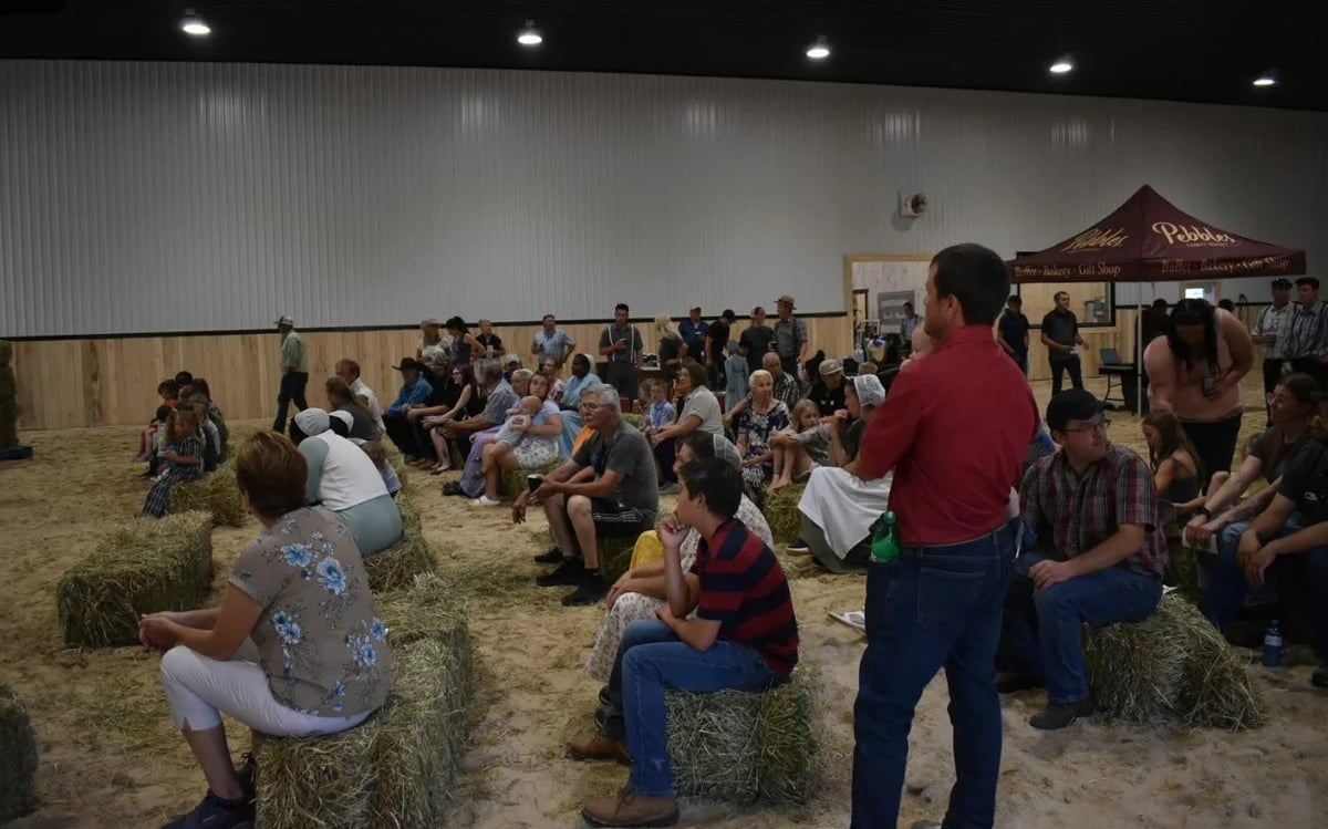 Large group of people sitting on hay bales in an indoor arena with livestock facilities and displays.