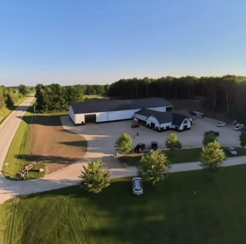Aerial view of a modern industrial building with solar panels, surrounded by green landscaping and parked vehicles