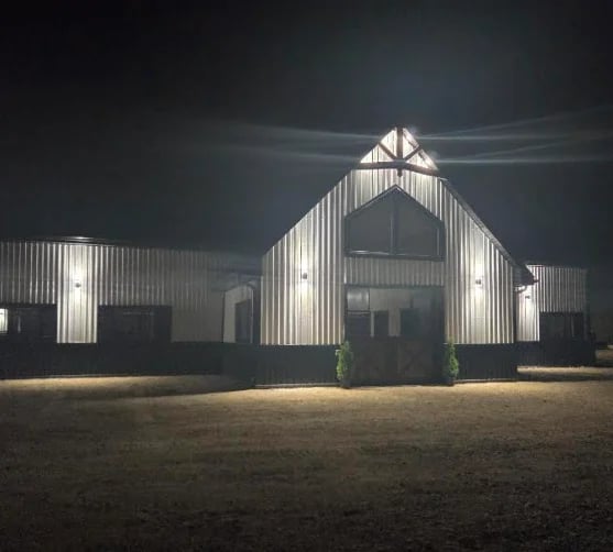 Modern white corrugated metal barn with peaked roof and entrance doors under dramatic dark sky, flanked by smaller outbuildings.